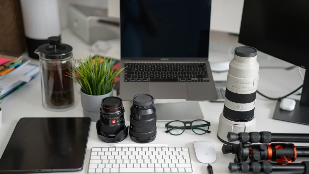 A photography workstation featuring a laptop, camera lenses, a tripod, and other equipment on a white desk.