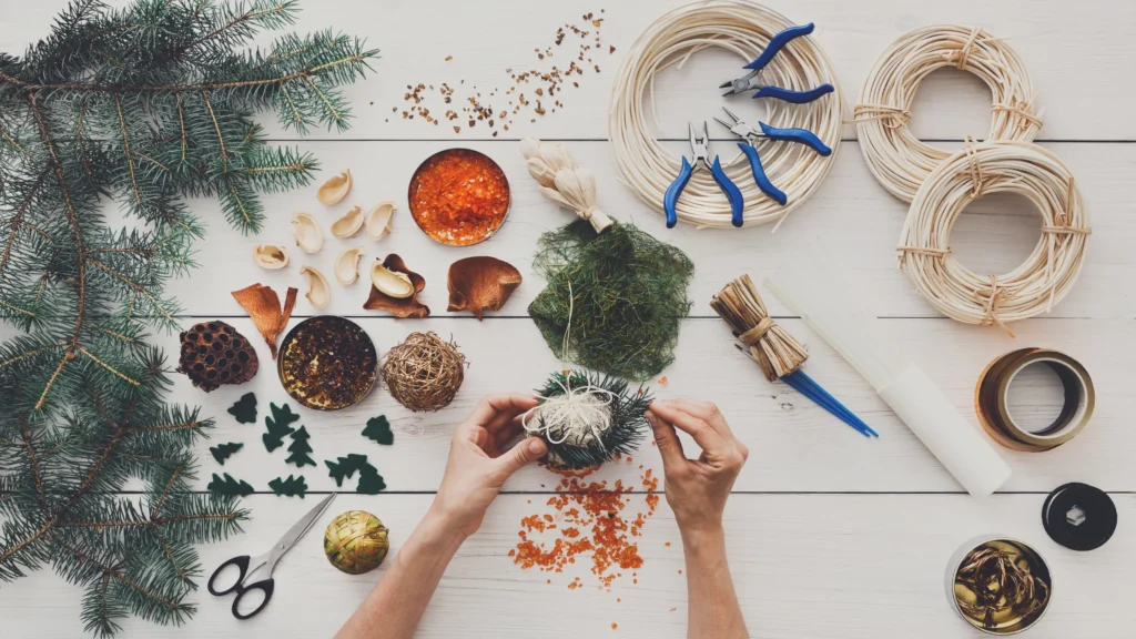 A person's hands arranging Christmas decorations on a white table, including red berries, pine cones, and ornaments, with scissors and twine also visible.







